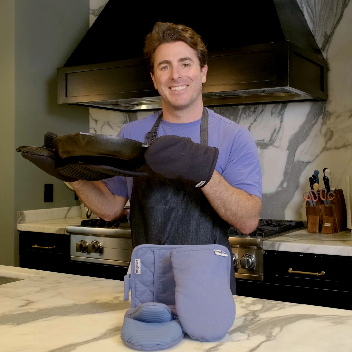 Man in a kitchen holding a cast iron pan with a blue hot pad on the bottom and a set of blue oven mitts and pot holder on the counter.