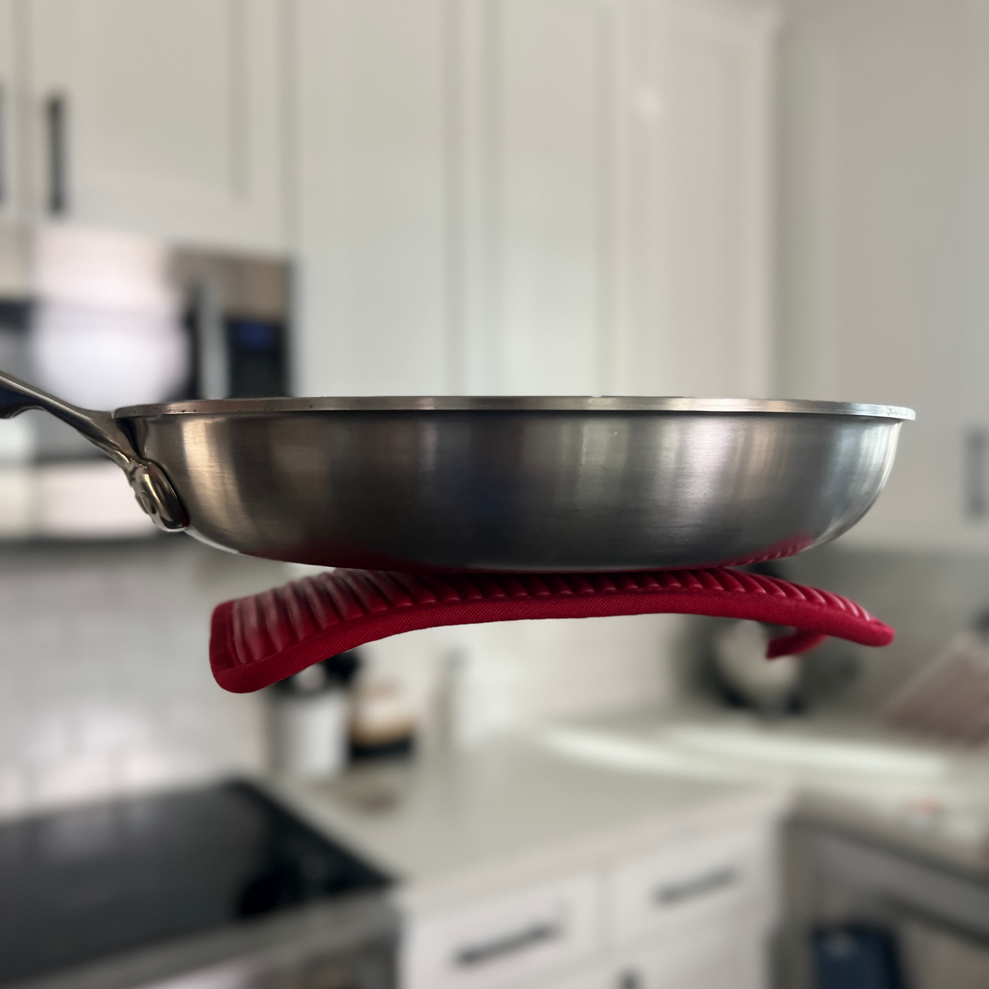 Stainless steel frying pan with red silicone handle holder on a blurred kitchen background