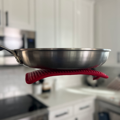 Stainless steel frying pan with red silicone handle holder on a blurred kitchen background