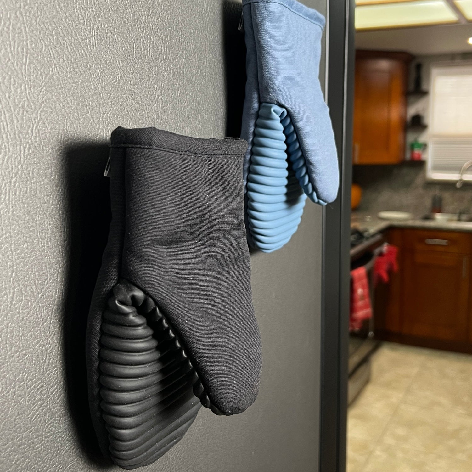 Black and blue oven mitts magnetized on a refrigerator side in a kitchen.