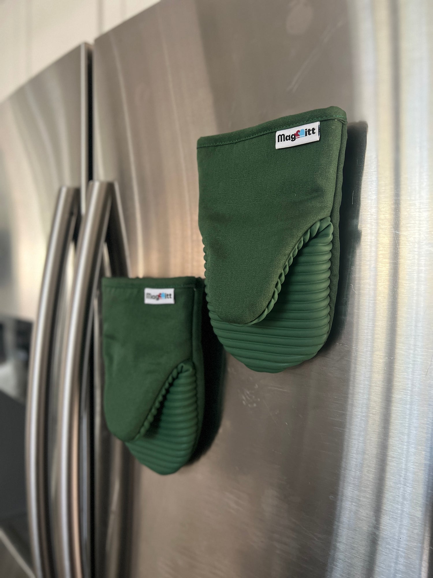 Green oven mitts and pot holder hanging on a stainless steel refrigerator.