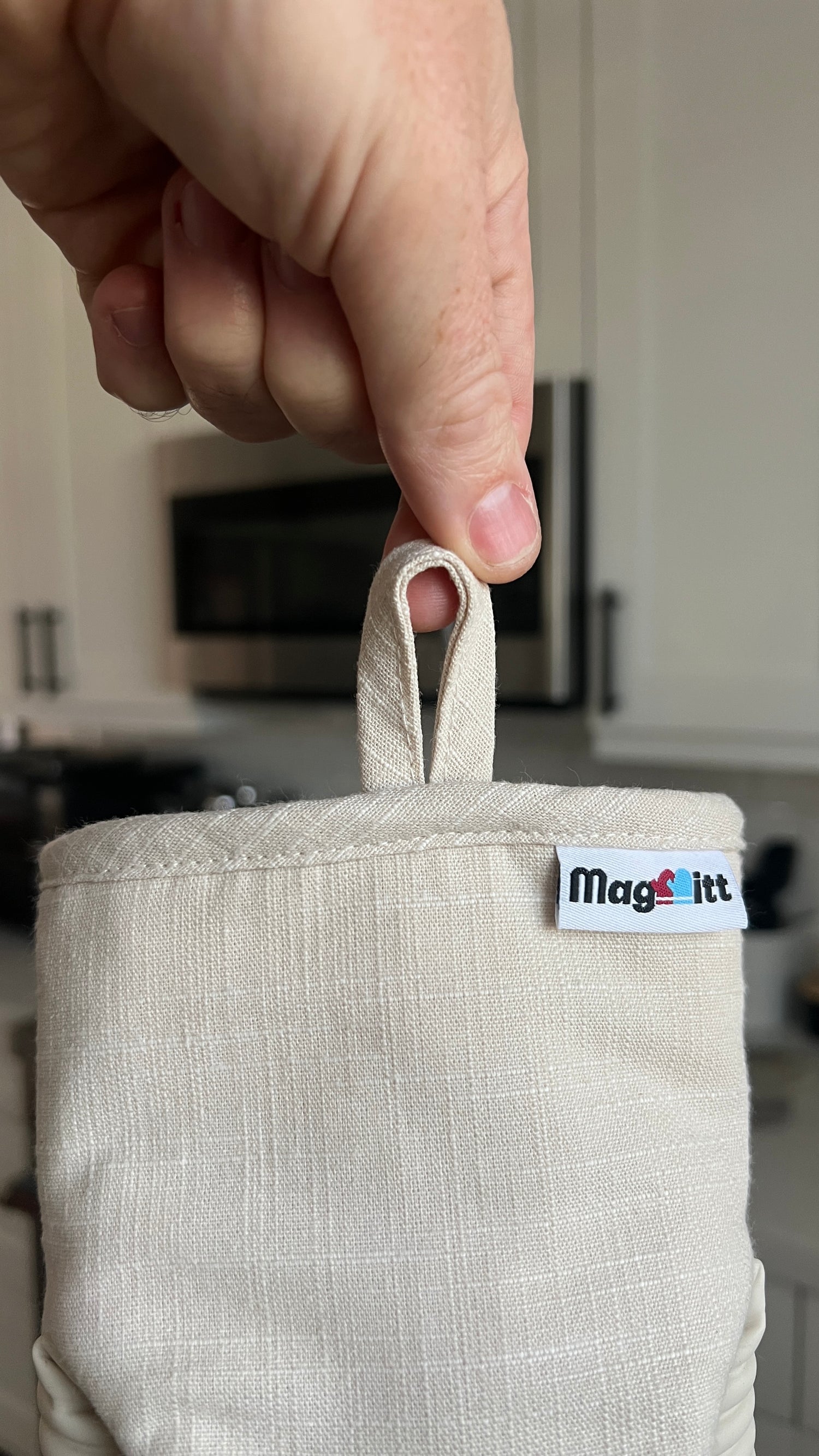Beige pot holder with 'MagMitt' logo held by a hand in a kitchen setting