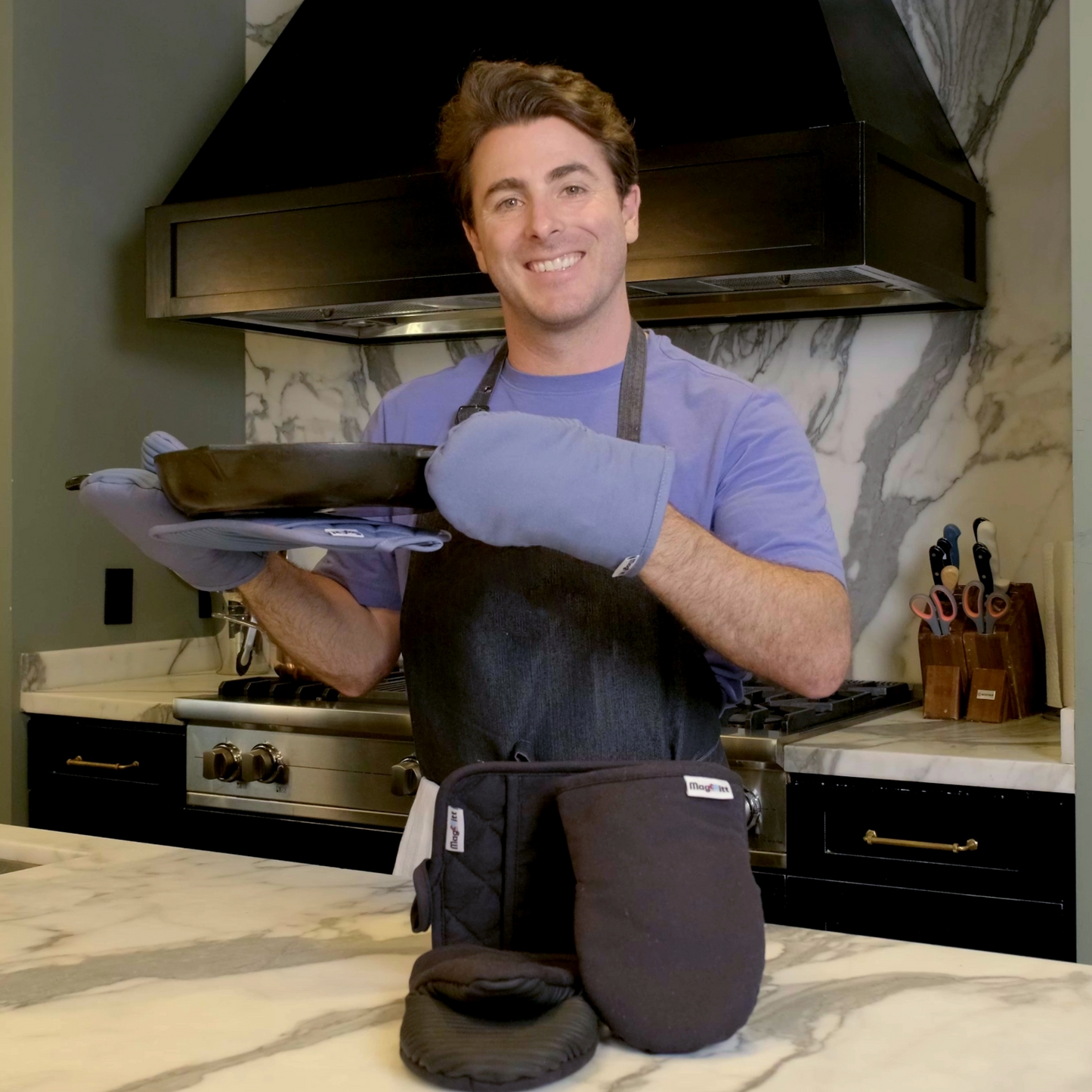Man in a kitchen wearing an apron and oven mitts, holding a pan.